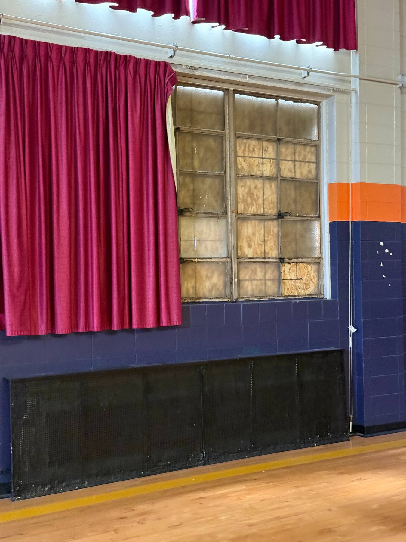 A vacant former classroom interior with a stage curtain, glass-block windows, and dated painted block walls.